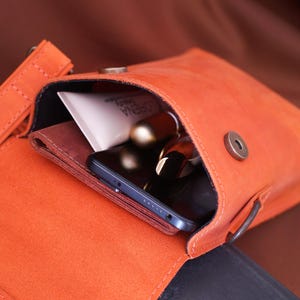 May include: An open, orange leather crossbody bag revealing its contents. Inside are a wallet, a smartphone, a tube of cream, and a lipstick. The bag has a magnetic clasp and a shoulder strap.