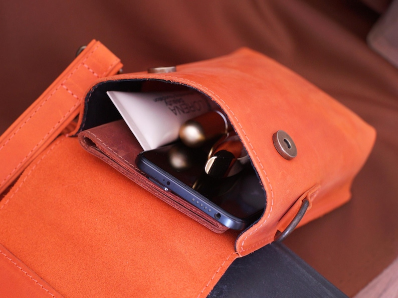May include: An open, orange leather crossbody bag revealing its contents. Inside are a wallet, a smartphone, a tube of cream, and a lipstick. The bag has a magnetic clasp and a shoulder strap.