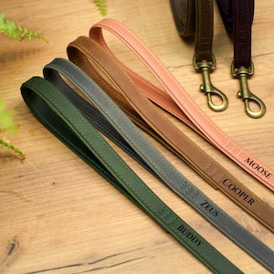 Puede incluir: Cinco correas de cuero para perros en diferentes colores con nombres personalizados: Buddy, Zeus, Cooper, Moose. Las correas están dispuestas en fila sobre una superficie de madera.