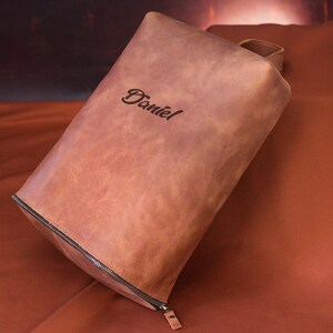 May include: A brown leather pouch with a zipper closure and a small handle. The name "Daniel" is embossed on the front. The pouch is rectangular and appears to be made of high-quality leather.