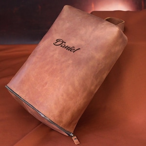 May include: A brown leather pouch with a zipper closure and a small handle. The name "Daniel" is elegantly embossed on the front. The pouch is rectangular and appears to be made of high-quality leather.