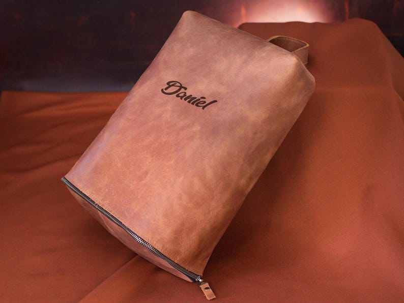 May include: A brown leather pouch with a zipper closure and a small handle. The name "Daniel" is elegantly embossed on the front. The pouch is rectangular and appears to be made of high-quality leather.