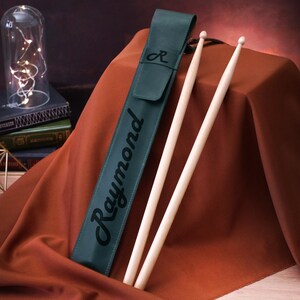 Puede incluir: Una funda de baquetas de cuero verde oscuro con el nombre "Raymond" impreso en ella. Dos baquetas de madera sobresalen de la funda.