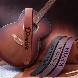 Puede incluir: Cuatro correas de guitarra de cuero marrón con diferentes nombres grabados: Dominie, Marshall, Randall y Kevin. Las correas están sobre una superficie marrón junto a una guitarra acústica de madera.