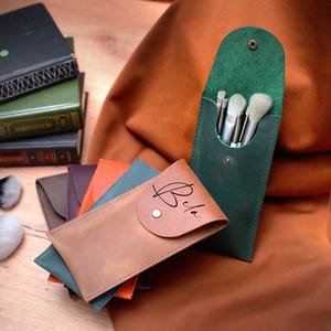 May include: Four leather brush holders, one green, one brown, one orange, and one blue. The brown holder has the word "Bria" written on it. The green holder has five brushes inside.