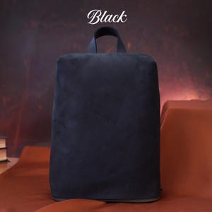 May include: A black backpack with a top handle, made of a soft, velvety material. The word "Black" is written in a script font at the top. The bag has a rectangular shape and a zipper at the bottom.