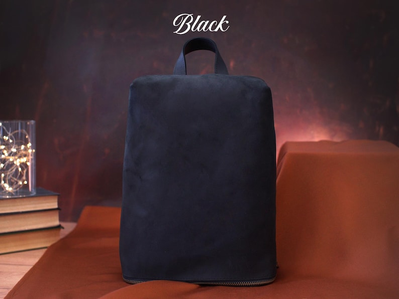 May include: A black backpack with a top handle, made of a soft, velvety material. The word "Black" is written in a script font at the top. The bag has a rectangular shape and a zipper at the bottom.