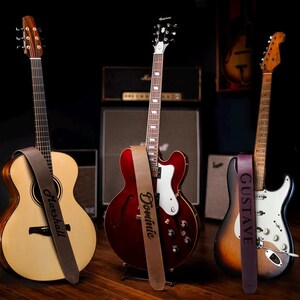 May include: Three guitars with personalized leather guitar straps. The straps are brown, burgundy, and dark purple. The guitars are a natural wood acoustic guitar, a red electric guitar, and a sunburst electric guitar.