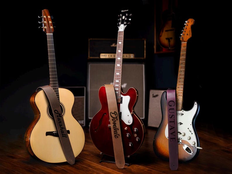 May include: Three guitars with personalized leather guitar straps. The straps are brown, burgundy, and dark purple. The guitars are a natural wood acoustic guitar, a red electric guitar, and a sunburst electric guitar.