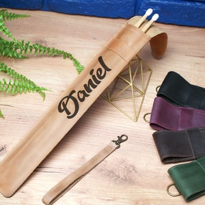 Pode incluir: Um porta-baquetas de couro marrom com o nome "Daniel" gravado nele. O porta-baquetas est&aacute; sobre uma superf&iacute;cie de madeira com um suporte de metal dourado e quatro outros porta-baquetas de couro em diferentes cores.
