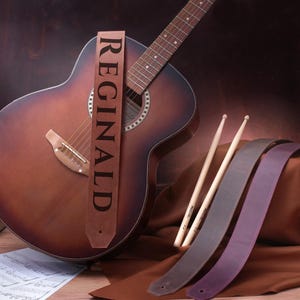 May include: A brown acoustic guitar with a leather guitar strap that says "REGINALD". The guitar is lying on a brown surface with two brown leather guitar straps and two wooden drumsticks.