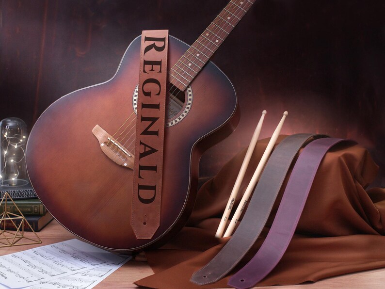 May include: A brown acoustic guitar with a leather guitar strap that says "REGINALD". The guitar is lying on a brown surface with two brown leather guitar straps and two wooden drumsticks.