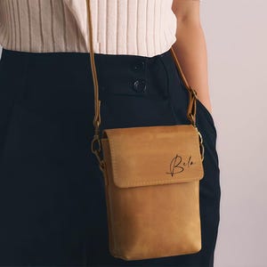May include: A tan leather crossbody bag with a flap closure and a long, thin strap. The bag has the name "Bela" written on the front in a cursive font. The bag is worn over the shoulder.