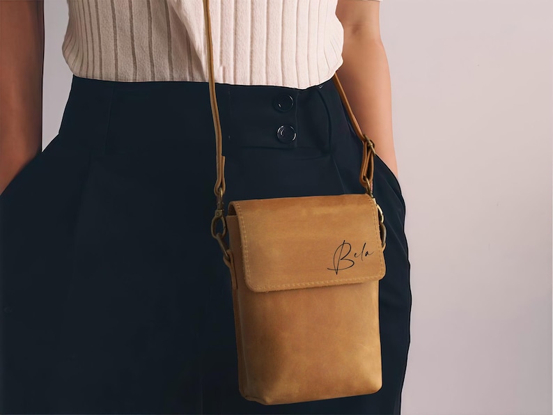 May include: A tan leather crossbody bag with a flap closure and a long, thin strap. The bag has the name "Bela" written on the front in a cursive font. The bag is worn over the shoulder.