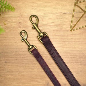 Puede incluir: Dos correas de cuero marr&oacute;n para perros con cierres de metal dorado. Las correas est&aacute;n sobre una superficie de madera.