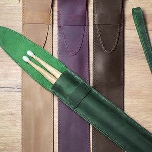 Pode incluir: Um conjunto de quatro estojos de couro para baquetas de bateria em bege, roxo, marrom e verde. O estojo verde tem um par de baquetas dentro. Os estojos t&ecirc;m uma al&ccedil;a e um la&ccedil;o para prender um chaveiro.