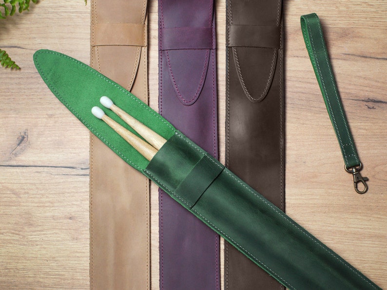 Pode incluir: Um conjunto de quatro estojos de couro para baquetas de bateria em bege, roxo, marrom e verde. O estojo verde tem um par de baquetas dentro. Os estojos t&ecirc;m uma al&ccedil;a e um la&ccedil;o para prender um chaveiro.