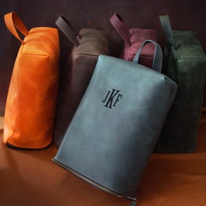 May include: A collection of leather bags in various colors, including orange, gray, brown, burgundy, and green. The gray bag in the foreground has the initials "JKF" embossed on it. Each bag has a handle.