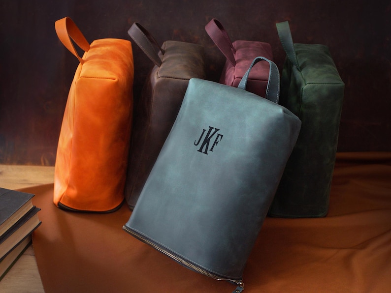 May include: A collection of leather bags in various colors, including orange, gray, brown, burgundy, and green. The gray bag in the foreground has the initials "JKF" embossed on it. Each bag has a handle.