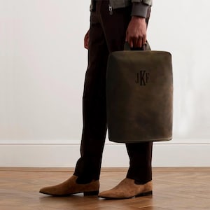 May include: A dark olive green leather tote bag with a handle, featuring the initials "JKF" in a darker shade. The bag is held by a person wearing brown pants and suede shoes.