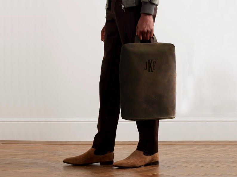 May include: A dark olive green leather tote bag with a handle, featuring the initials "JKF" in a darker shade. The bag is held by a person wearing brown pants and suede shoes.
