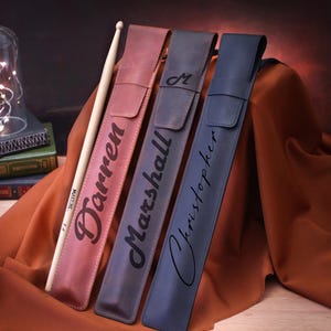 Puede incluir: Tres estuches para baquetas personalizados en cuero marr&oacute;n, azul y azul oscuro. Los estuches est&aacute;n grabados con los nombres "Darren", "Marshall" y "Christopher".
