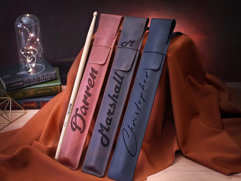 Puede incluir: Tres estuches para baquetas personalizados en cuero marr&oacute;n, azul y azul oscuro. Los estuches est&aacute;n grabados con los nombres "Darren", "Marshall" y "Christopher".