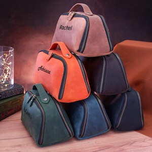 May include: Six personalised leather travel bags stacked on top of each other. The bags are in various colours, including brown, orange, blue, and green. Each bag has a zipper closure and a handle. The bags are personalised with the names "Rachel", "Adriana", "Agnes", and other names.