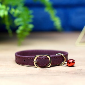 K&ouml;nnte beinhalten: Ein burgunderfarbenes Lederhalsband f&uuml;r Katzen mit einer goldenen Schnalle und einem roten Gl&ouml;ckchen-Anh&auml;nger.