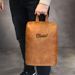 May include: A rectangular, tan leather bag with a handle is held by a person. The bag has the name "Daniel" embossed on the front. The bag is a light brown color and appears to be made of high-quality leather.