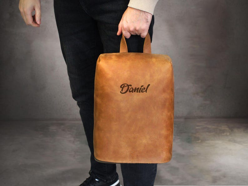 May include: A rectangular, tan leather bag with a handle is held by a person. The bag has the name "Daniel" embossed on the front. The bag is a light brown color and appears to be made of high-quality leather.