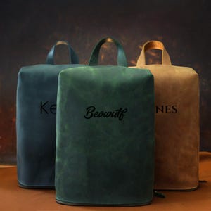May include: Three leather bags in shades of teal, green, and tan, each with a handle and a zipper. The bags have personalized text: "Ke", "Beowulf", and "NES". The bags are rectangular with rounded tops.