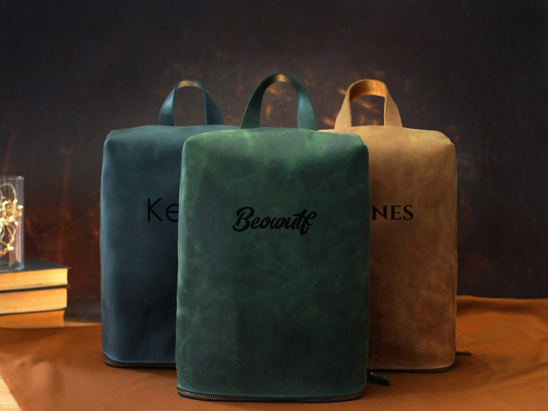 May include: Three leather bags in shades of teal, green, and tan, each with a handle and a zipper. The bags have personalized text: "Ke", "Beowulf", and "NES". The bags are rectangular with rounded tops.