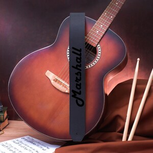 May include: A black leather guitar strap with the word "Marshall" printed in white lettering. The strap is attached to a brown acoustic guitar.