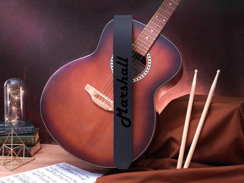 May include: A black leather guitar strap with the word "Marshall" printed in white lettering. The strap is attached to a brown acoustic guitar.