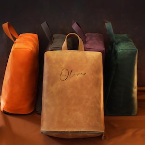 May include: A collection of leather tote bags in various colors, including orange, brown, dark green, and purple. The bags have a rectangular shape with a handle and a zipper at the bottom. The word "Oliver" is written on the front of one bag.