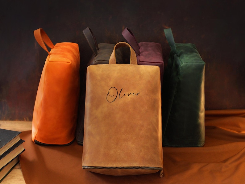 May include: A collection of leather tote bags in various colors, including orange, brown, dark green, and purple. The bags have a rectangular shape with a handle and a zipper at the bottom. The word "Oliver" is written on the front of one bag.