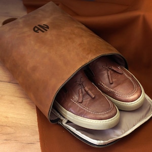 以下が含まれることがあります： 茶色の革製の靴バッグ。ジッパー付きで、白いソールが付いた茶色の革靴が1足入っています。バッグにはハンドルがあり、前面にはイニシャル「AN」がエンボス加工されています。