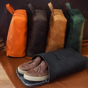May include: A collection of leather shoe bags in various colors: orange, dark brown, tan, and green. A black shoe bag with the word "Marshall" is open, revealing a pair of brown leather shoes with white soles. The bags have a handle.