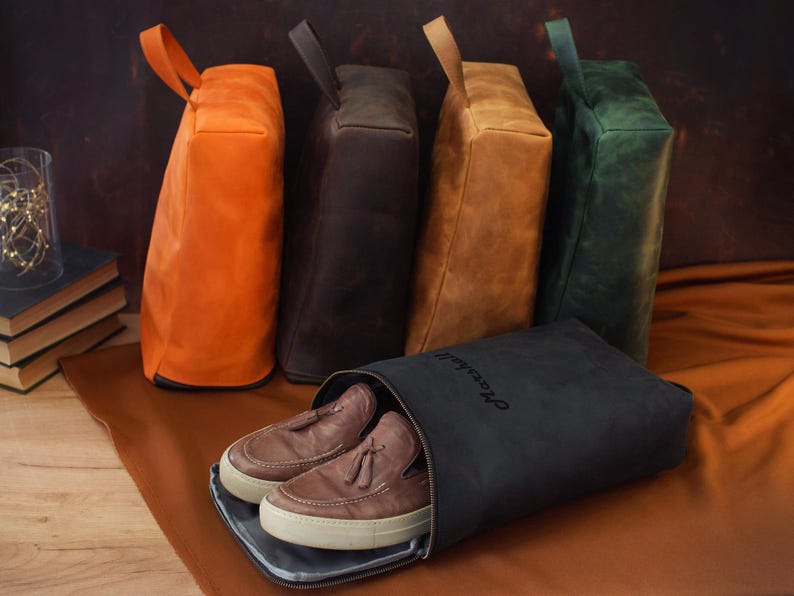 May include: A collection of leather shoe bags in various colors: orange, dark brown, tan, and green. A black shoe bag with the word "Marshall" is open, revealing a pair of brown leather shoes with white soles. The bags have a handle.