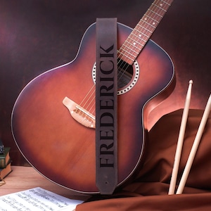 May include: A brown leather guitar strap with the name "FREDERICK" embossed in white. The strap is hanging on a brown acoustic guitar with a natural wood finish. The guitar is resting on a brown fabric surface.