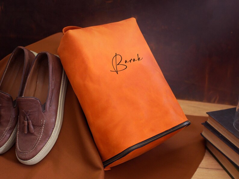 May include: An orange leather shoe bag with a black zipper and the name "Barak" in script. Brown leather loafers with tassels are next to the bag. The bag is on a brown surface.