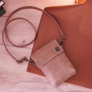 May include: A light brown leather crossbody bag with a flap closure and a long strap. The bag has the initials "JKF" embossed on the front. The bag is lying on a wooden surface next to a rust-colored fabric.