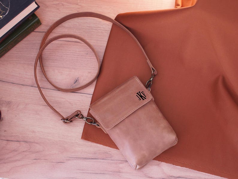 May include: A light brown leather crossbody bag with a flap closure and a long strap. The bag has the initials "JKF" embossed on the front. The bag is lying on a wooden surface next to a rust-colored fabric.