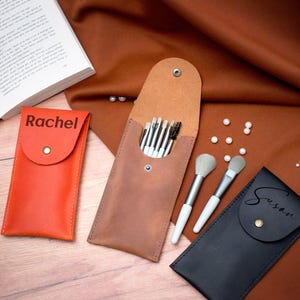 May include: Three personalized leather makeup brush holders. The holders are brown, orange, and black. The brown holder has a set of makeup brushes inside. The orange holder has the name "Rachel" on it. The black holder has the name "Susan" on it.
