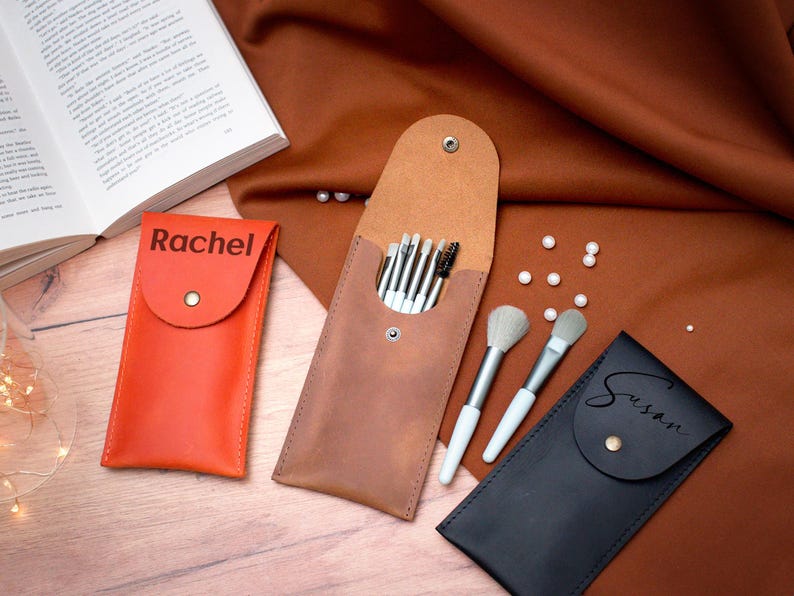 May include: Three personalized leather makeup brush holders. The holders are brown, orange, and black. The brown holder has a set of makeup brushes inside. The orange holder has the name "Rachel" on it. The black holder has the name "Susan" on it.