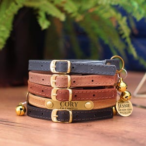 May include: Five leather cat collars in different colors, stacked on top of each other. The top collar is navy blue, the next is light brown, then dark brown, then light beige, and the bottom collar is black. The beige collar has a gold name tag that reads "Cory I'm chipped". A gold bell charm with a gold tag that reads "Jessie 121-654-1254" is attached to the bottom collar.