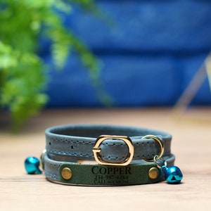 K&ouml;nnte beinhalten: Ein blaues Lederhalsband f&uuml;r Katzen mit einer goldenen Schnalle und einem gr&uuml;nen Lederanh&auml;nger mit der Aufschrift "COPPER 234-987-4281 CALL MY MOM". Das Halsband hat zwei blaue Gl&ouml;ckchen.
