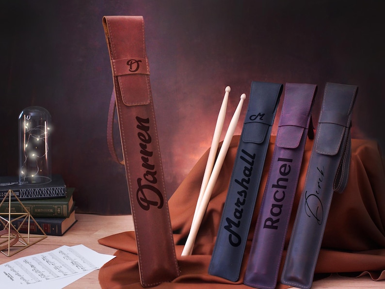 Puede incluir: Cuatro estuches de baquetas de cuero personalizados con los nombres Darren, Marshall, Rachel y Derek. Los estuches son marrones y morados. Las baquetas son de madera y tienen un acabado natural.