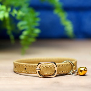 K&ouml;nnte beinhalten: Ein braunes Lederhalsband f&uuml;r Katzen mit einer goldenen Schnalle und einem goldenen Gl&ouml;ckchen-Anh&auml;nger.